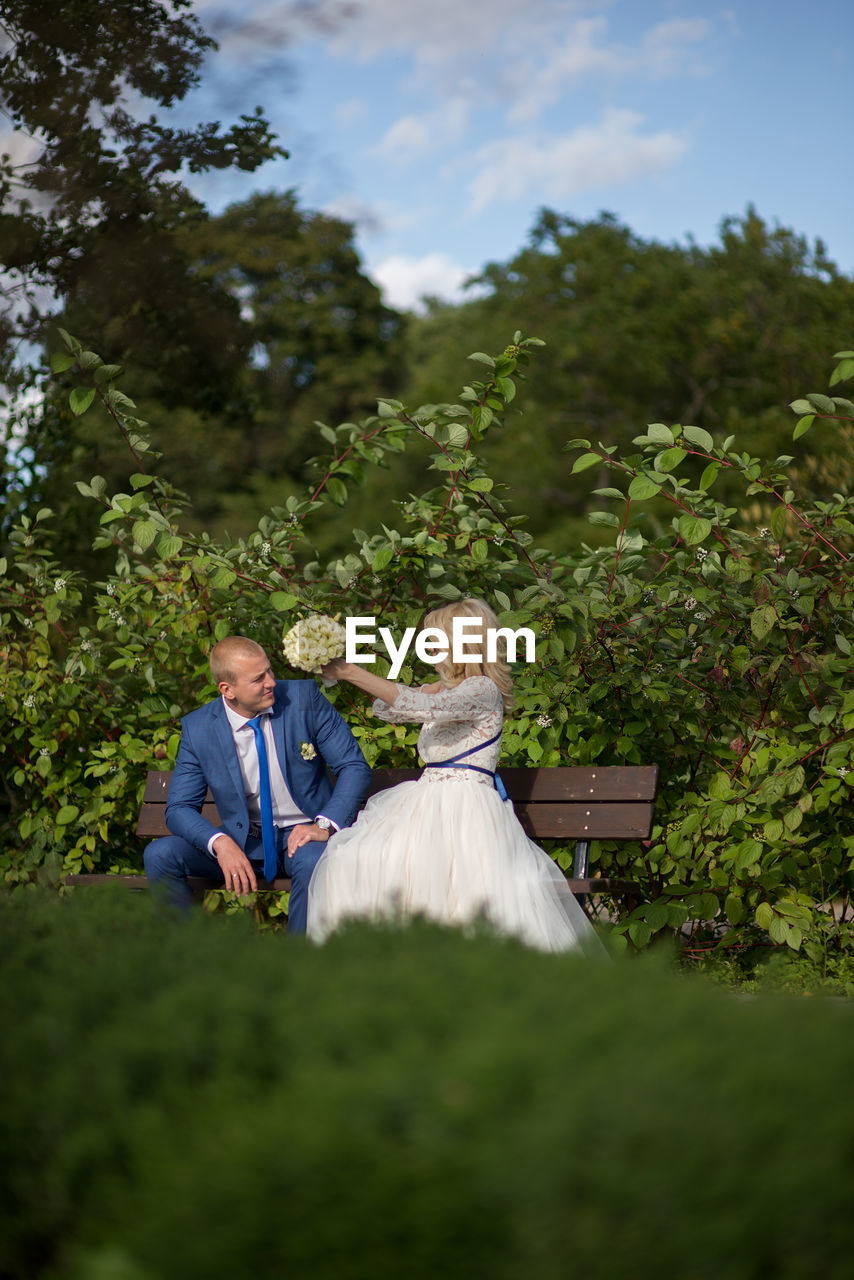 Bridal couple sitting on bench against plants at park