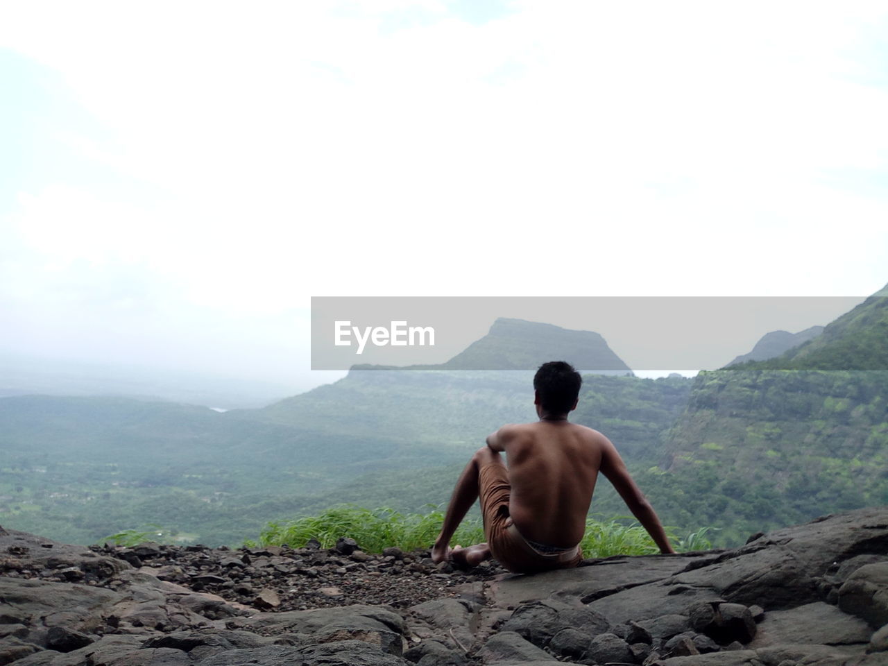 Rear view of shirtless man sitting on rock against mountains
