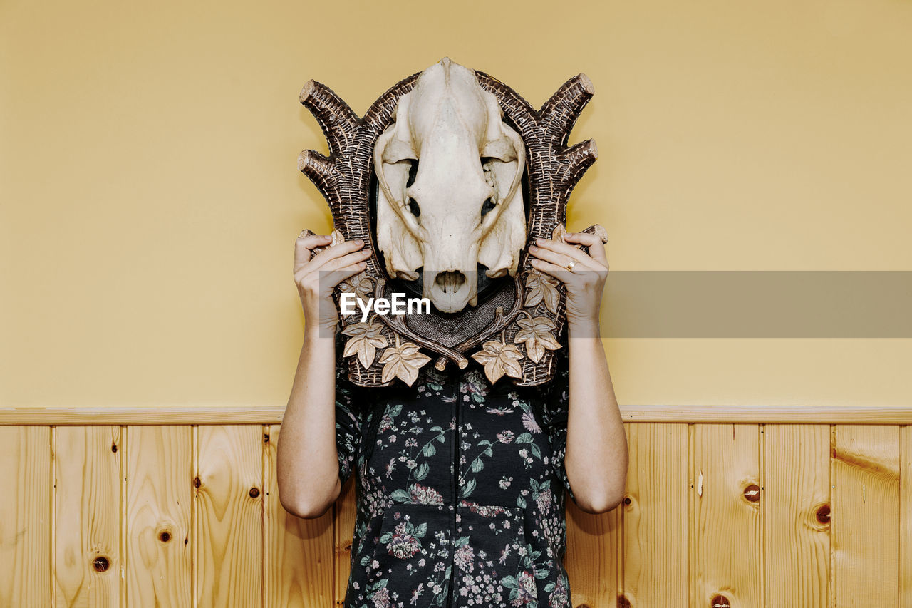 Woman with face covered by animal skull standing against wall