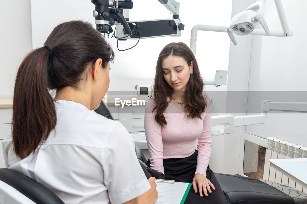 Female dentist filling in papers while talking to young patient in dental office