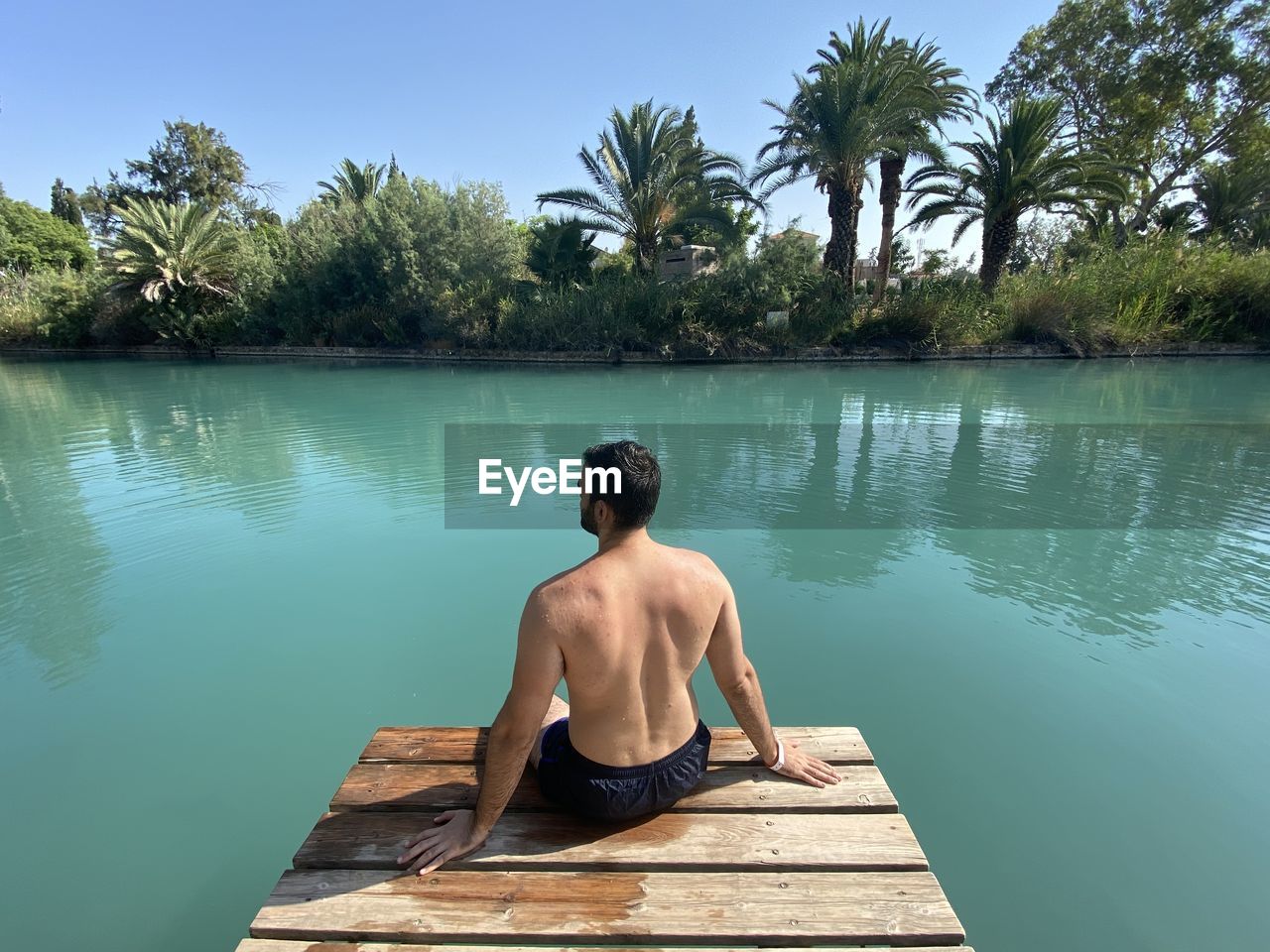 REAR VIEW OF SHIRTLESS MAN SITTING AT SWIMMING POOL