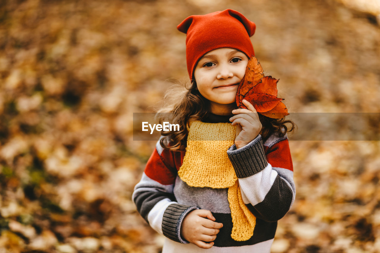 Portrait of a little girl a child in a warm hat walk holding an autumn leaf in fall forest outdoor