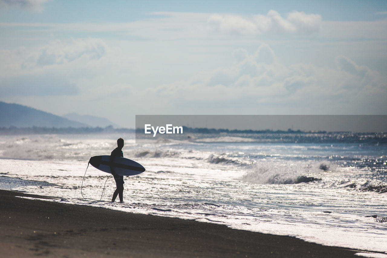 Surfer standing on beach by sea against sky