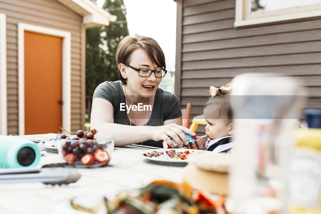 Mother feeding daughter while sitting at table in backyard