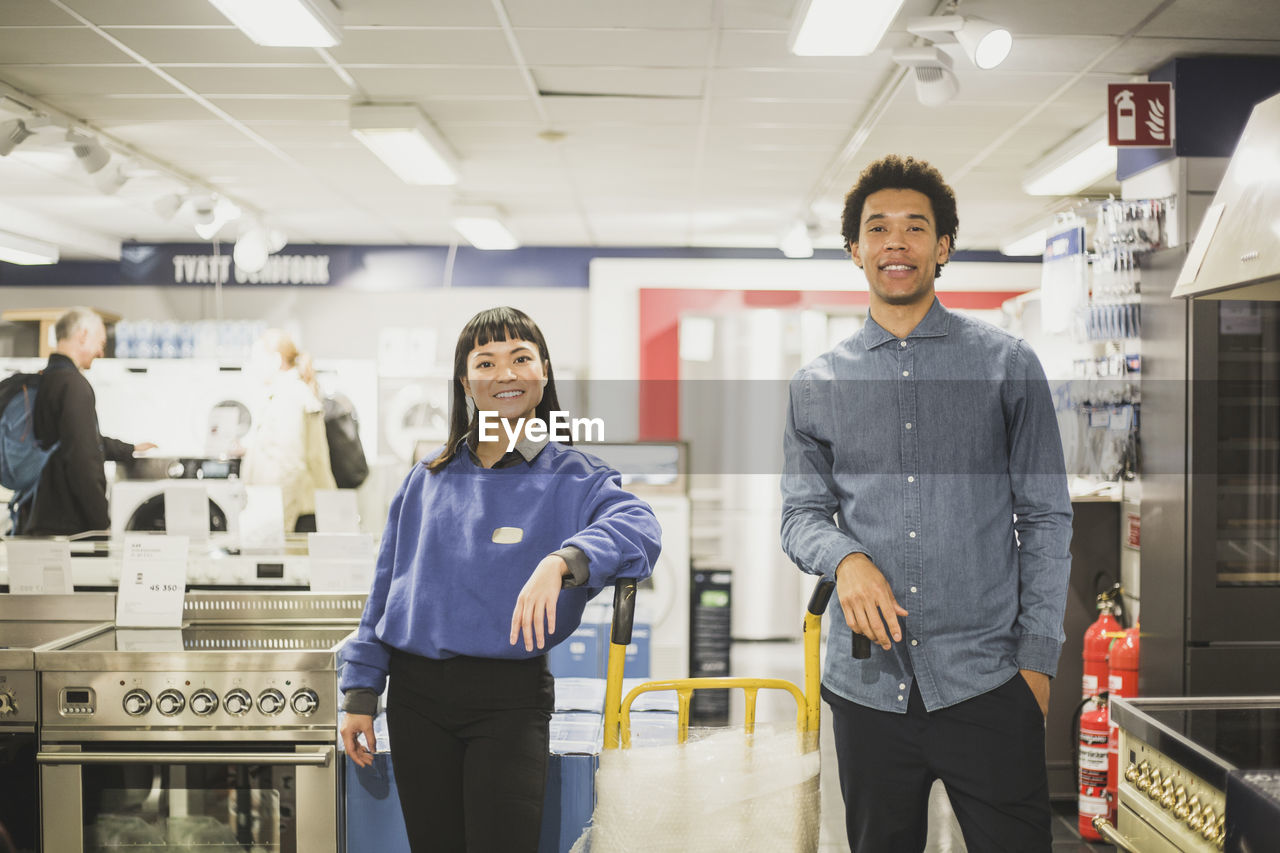 Portrait of smiling salesman and saleswoman standing by luggage cart in electronics store