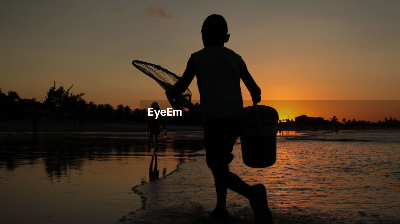 Silhouette boy with bucket and fishing net walking at beach during sunset