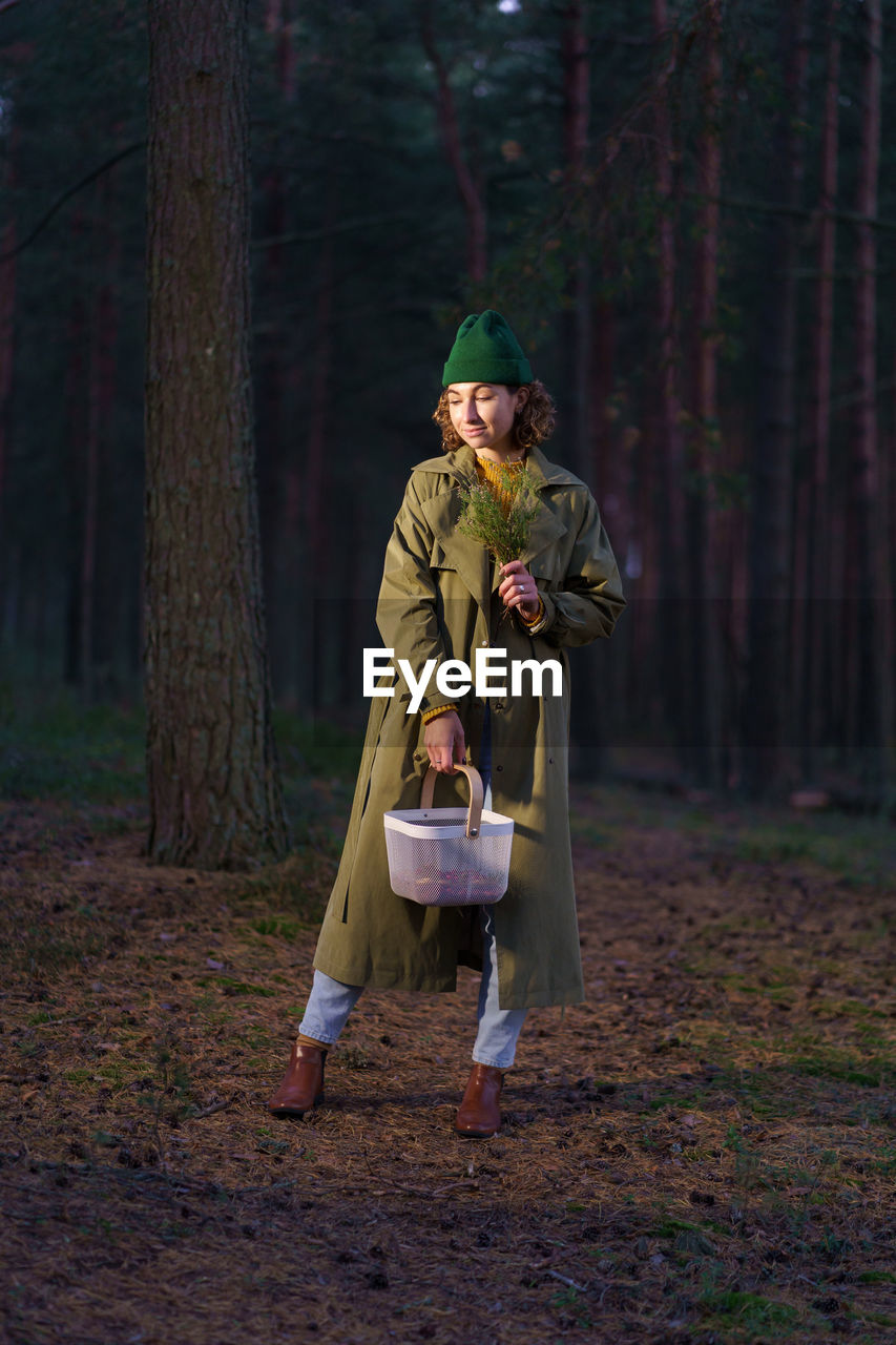 Young woman in hipster hat and trendy coat smiles standing with basket at dark trees