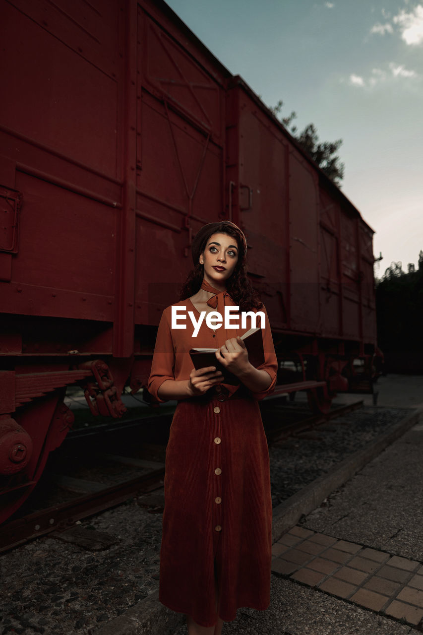 Dark haired female totally wearing terracotta clothes in vintage style standing near terracotta car train and holding open book in hands having dreaming look