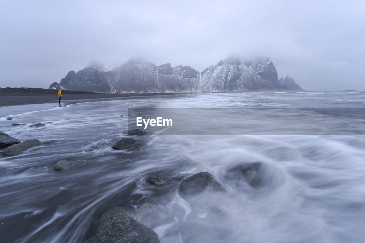 Distant view of unrecognizable traveler standing on wet black sand beach near waving sea against mountain ridge and cloudy sky in iceland