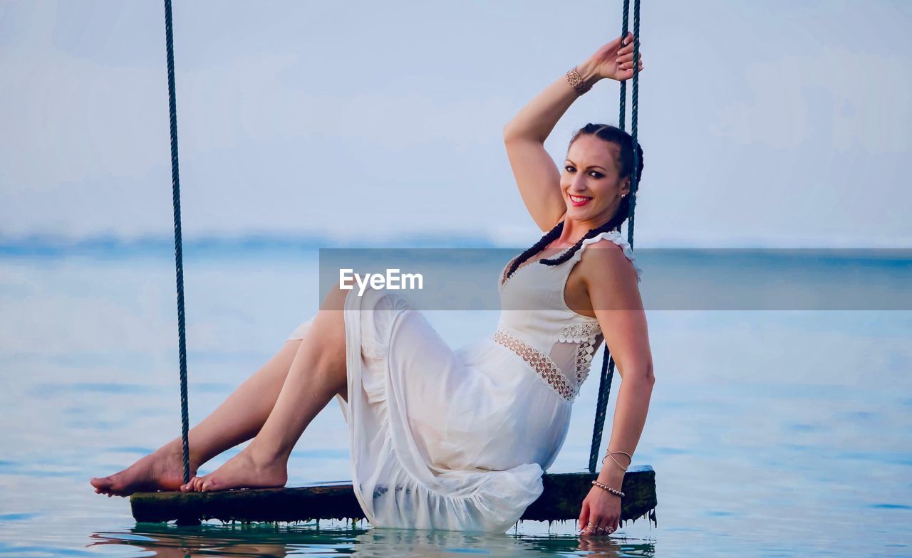 Full length portrait of smiling woman sitting on swing at sea