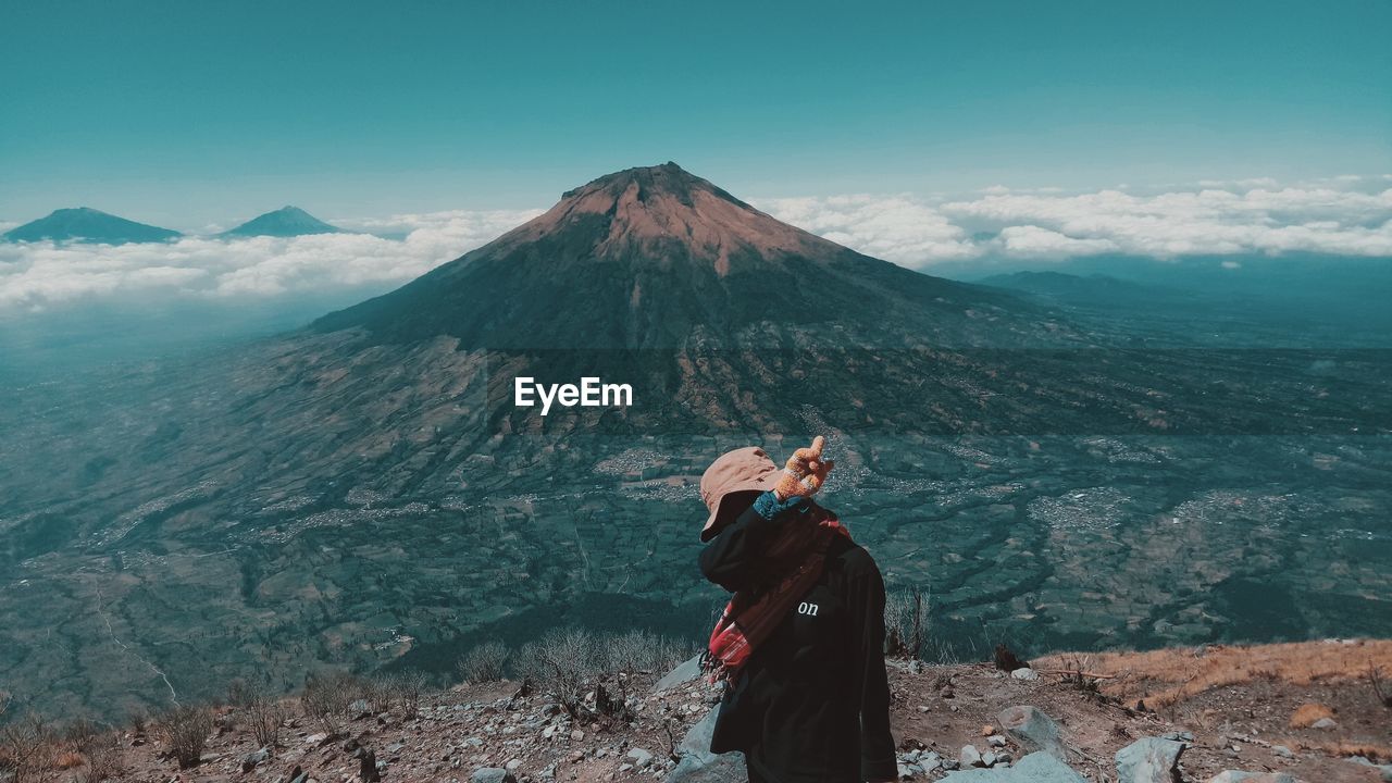 REAR VIEW OF PERSON STANDING ON MOUNTAIN AGAINST SKY