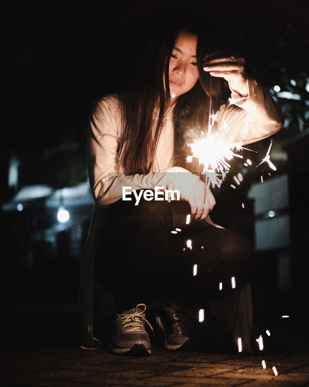 Young woman holding illuminated sparkler while crouching at night
