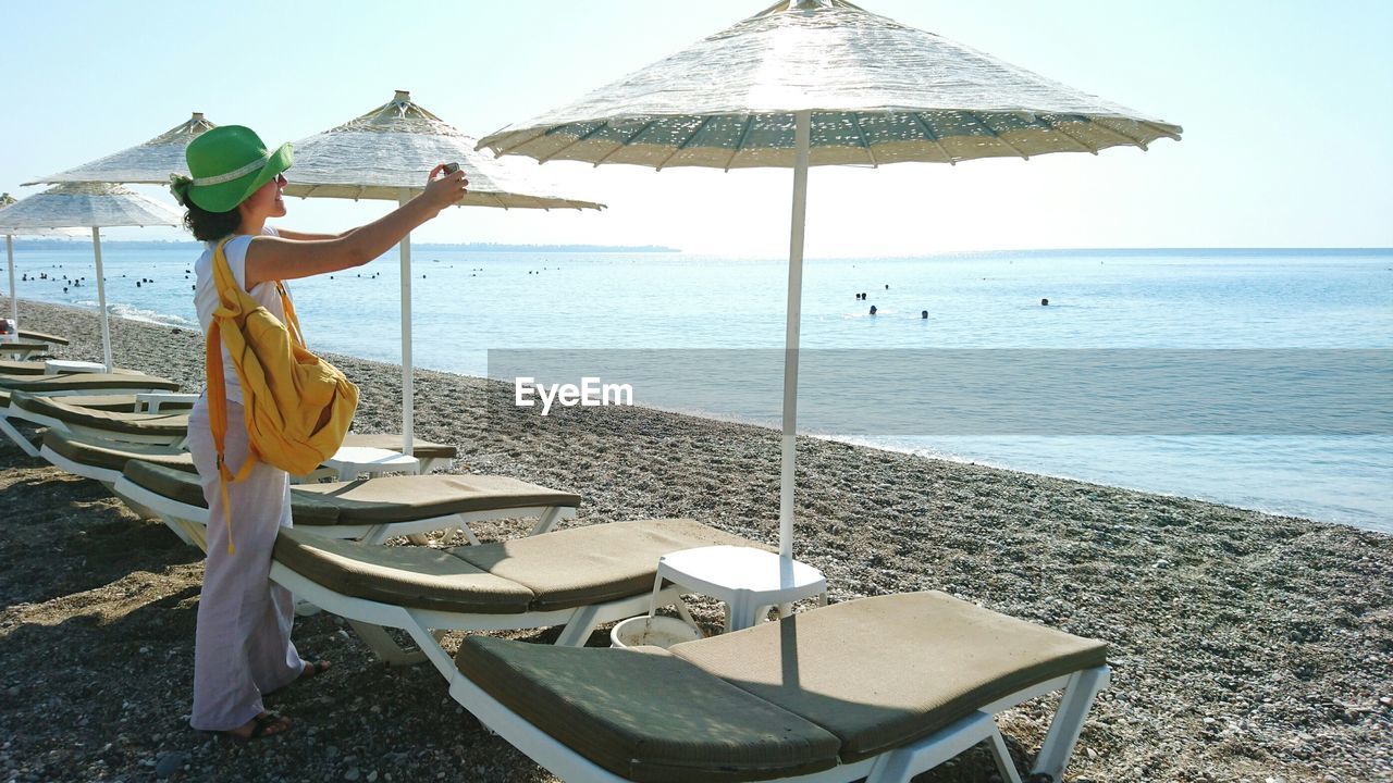 Woman photographing with smart phone while standing by parasols and lounge chairs at beach