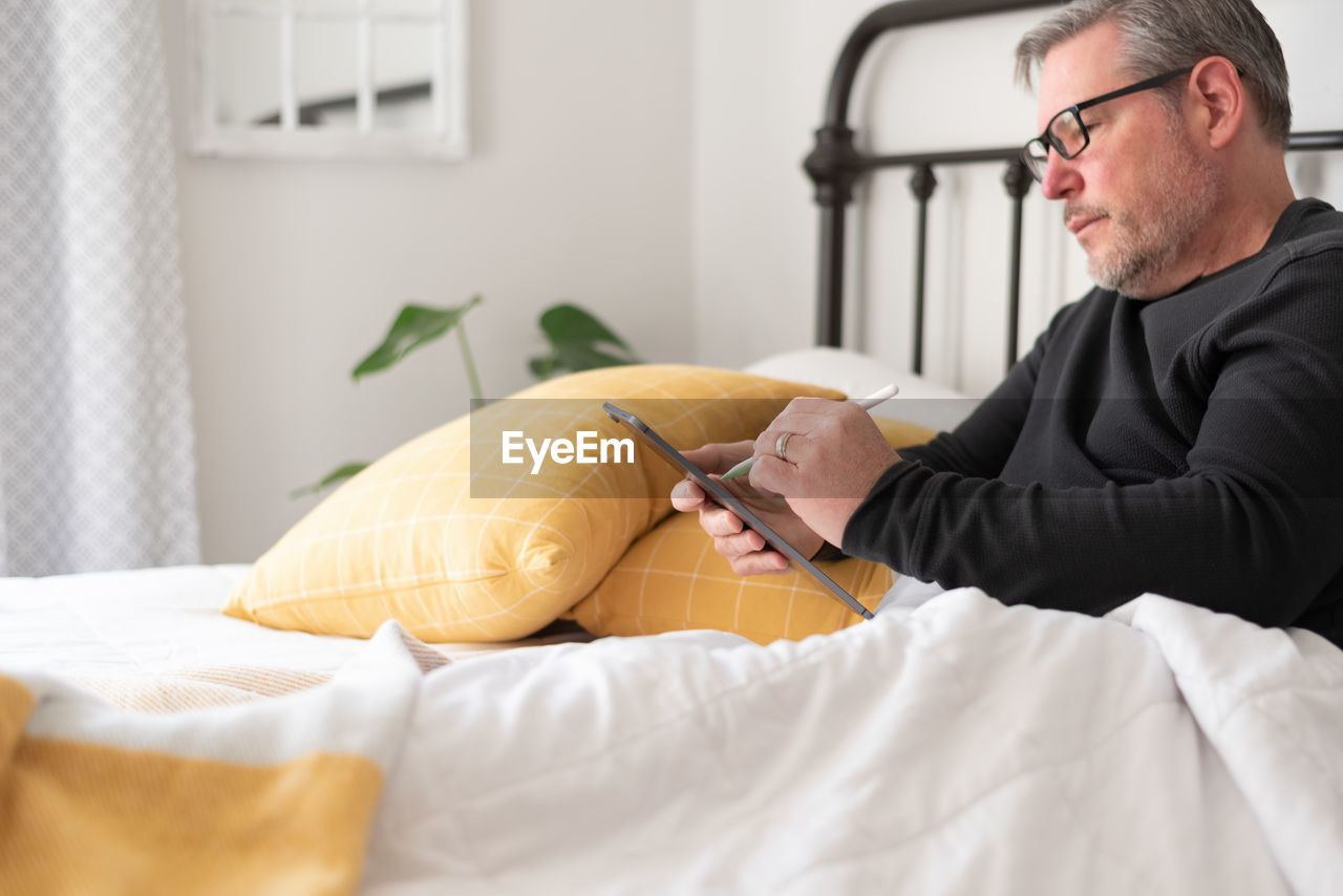 Mature man sitting in bed using a stylus pen and digital tablet