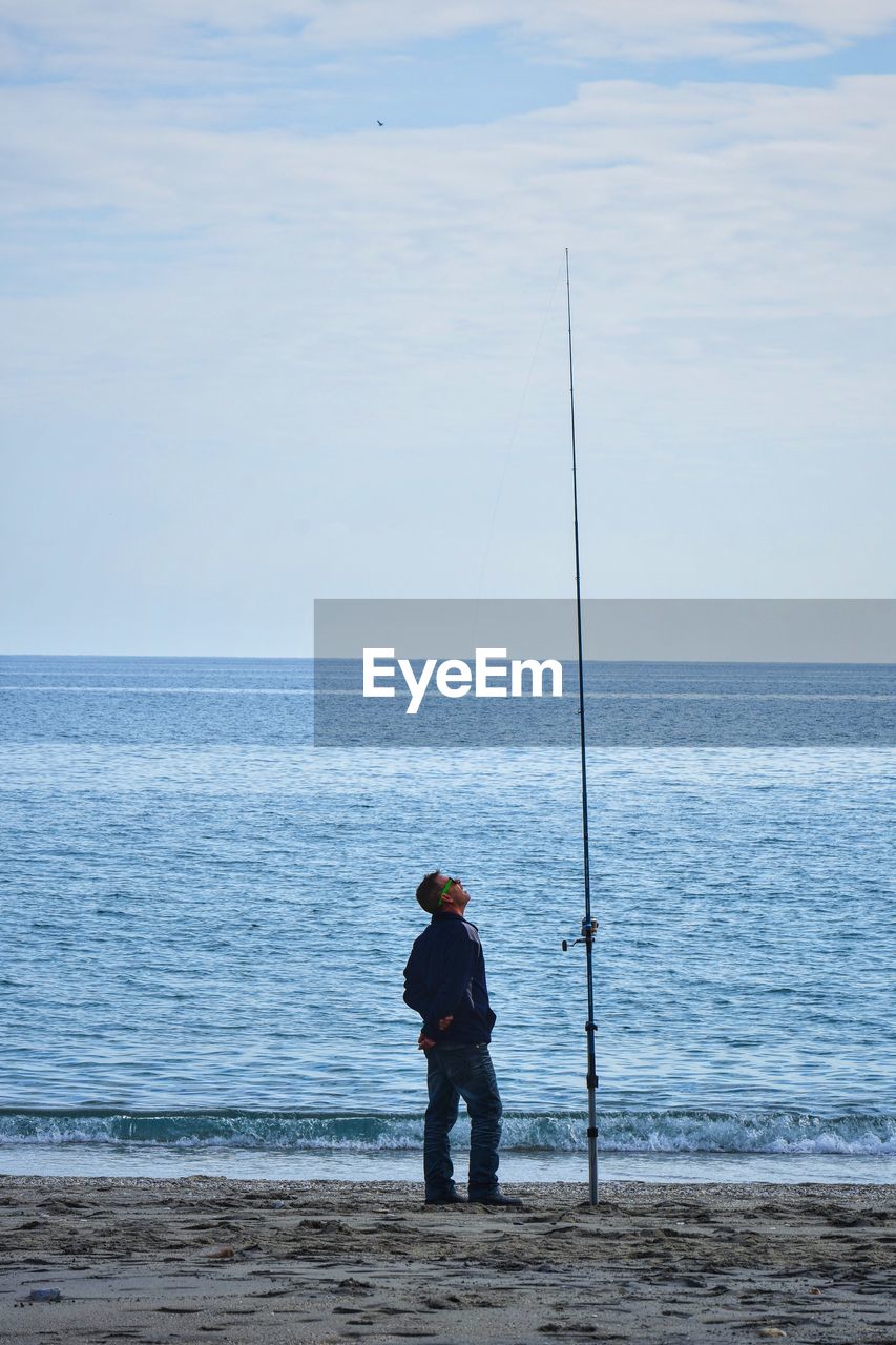 Full length of man looking at fishing rod at beach against sky