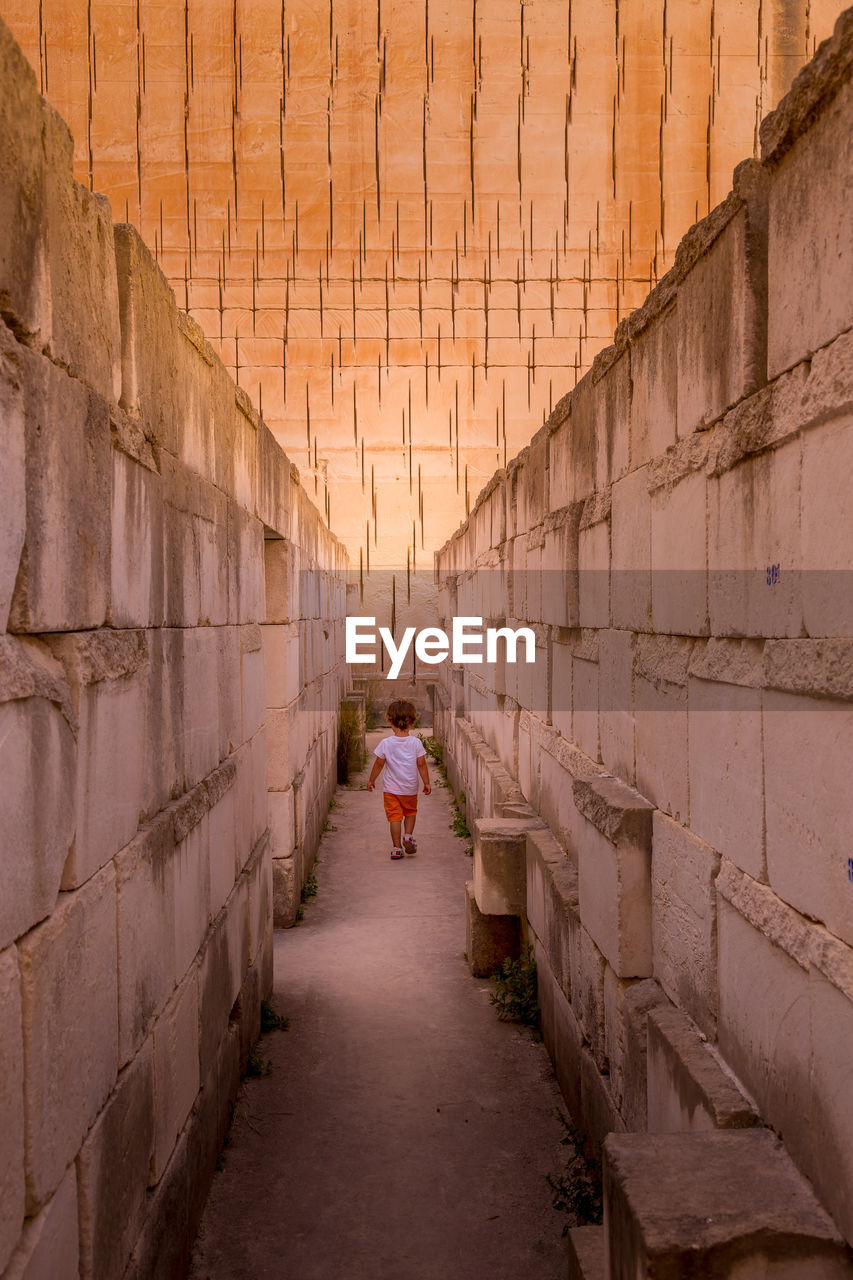 REAR VIEW OF WOMAN WALKING ON NARROW ALLEY AMIDST BUILDINGS