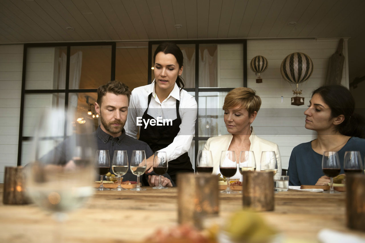 Woman wearing apron talking to business people while pointing at wineglass on table
