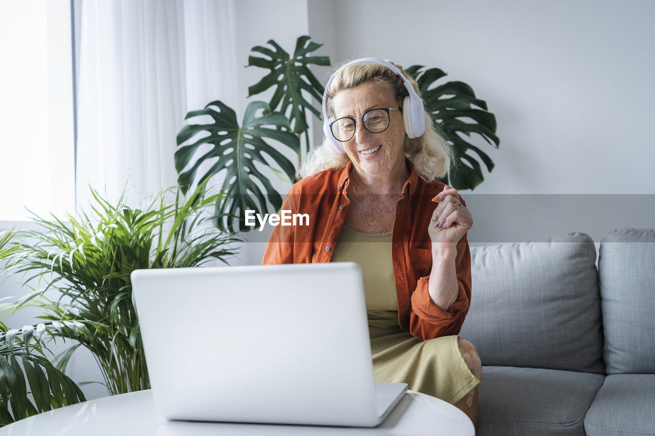 Smiling woman enjoying music through headphones and laptop sitting at home