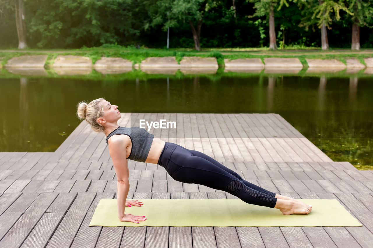 A slim woman on a wooden platform by a pond in a park in summer, does yoga