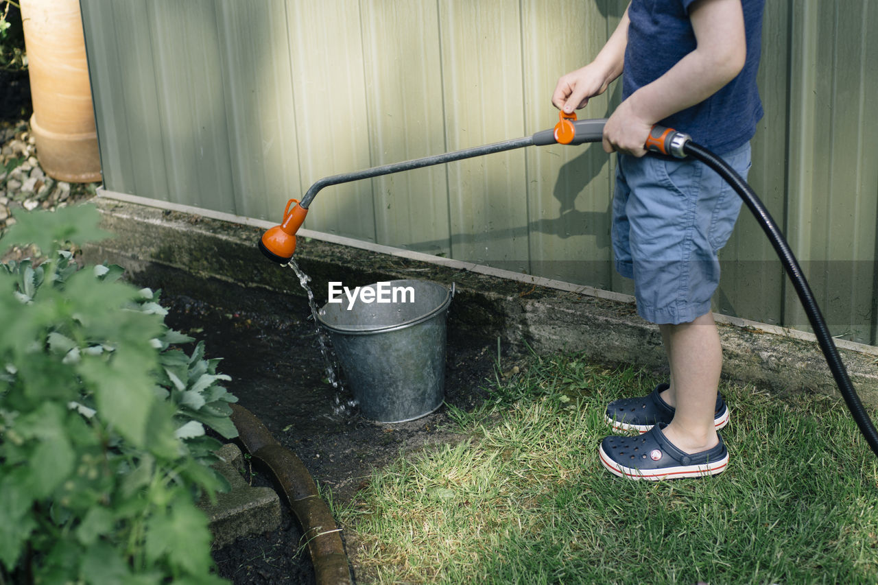 Low section of boy pouring water with garden hose in bucket at yard