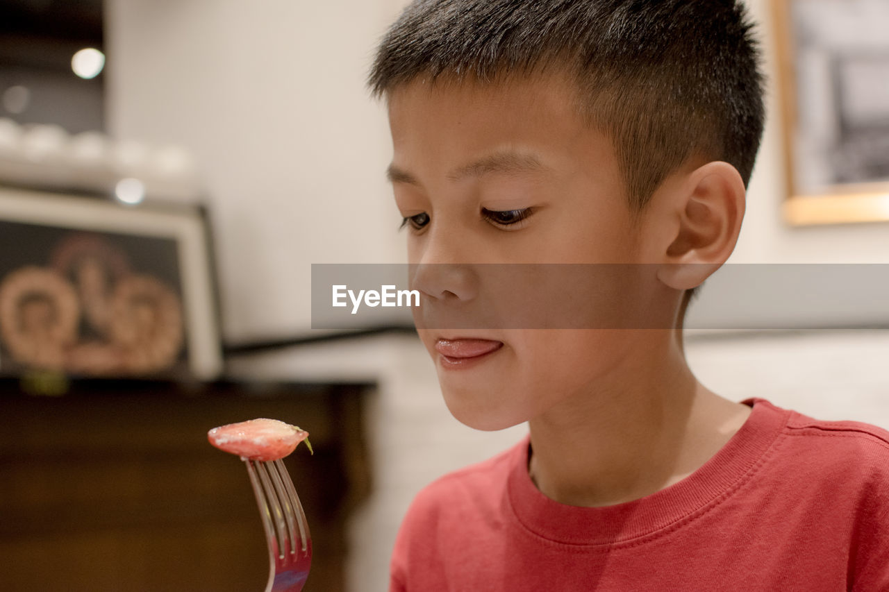 Boy sticking out tongue while looking at strawberry slice in fork at home