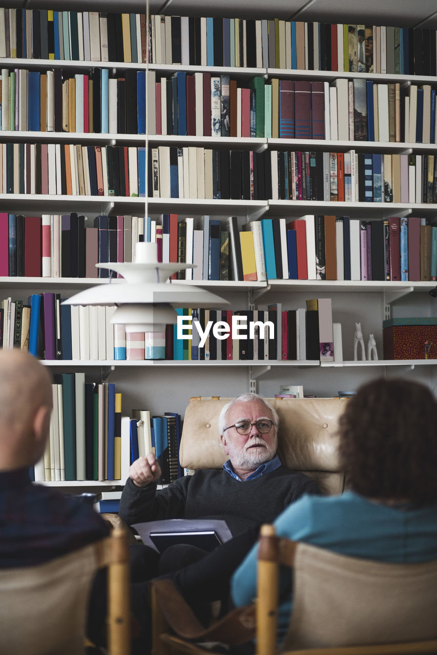 Senior therapist explaining couple while sitting against bookshelf