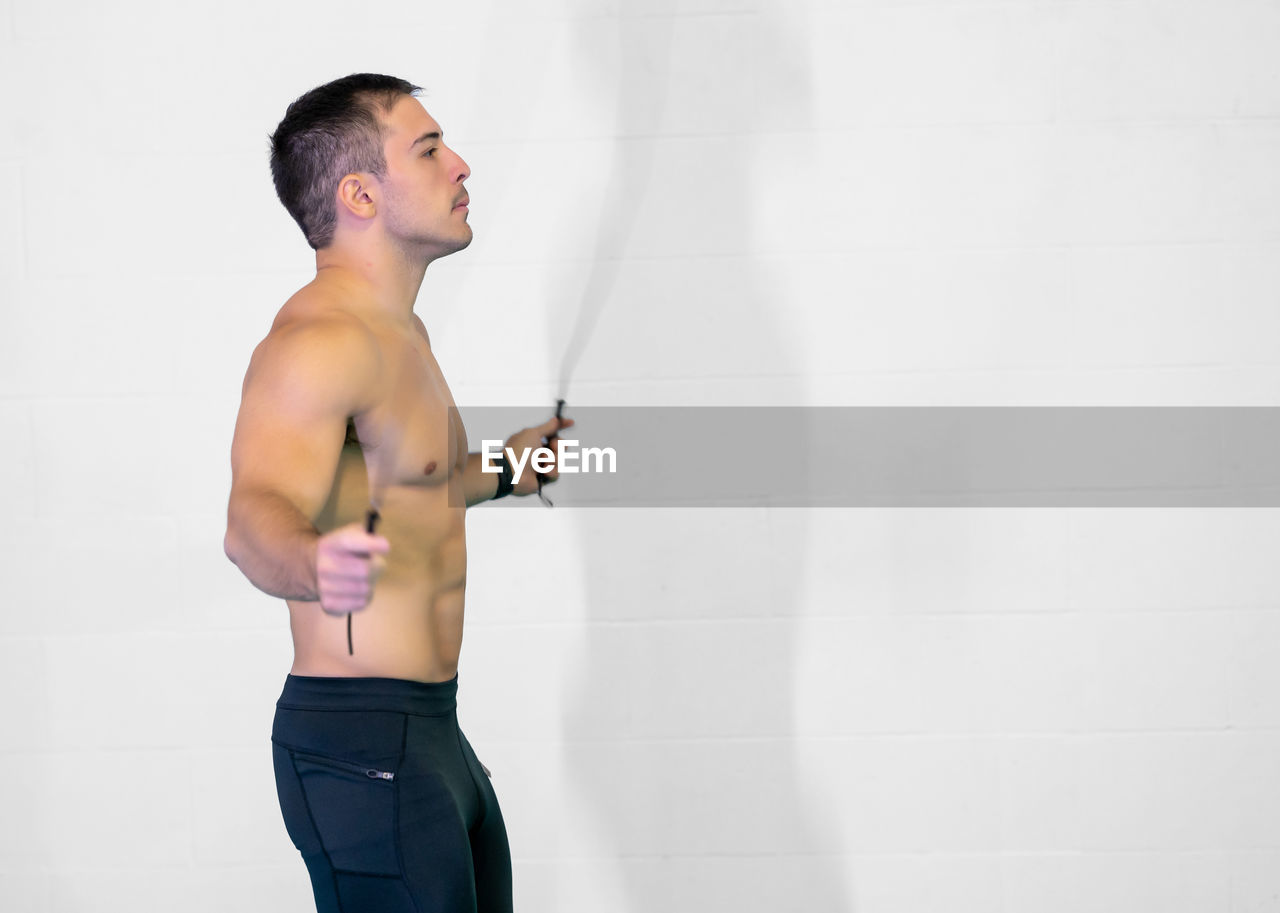 one person, lifestyles, copy space, adult, standing, limb, wall - building feature, exercising, young adult, indoors, arm, photo shoot, side view, men, sports, muscular build, trunk, athlete, sports clothing, looking, strength, wellbeing, three quarter length, clothing, sports training, white, vitality, waist up, person
