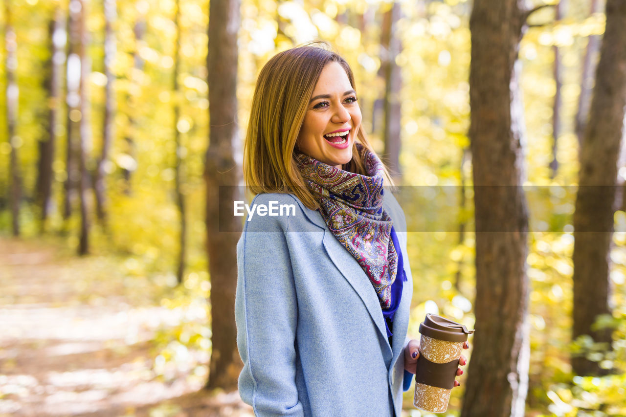 SMILING YOUNG WOMAN STANDING AGAINST TREES AT FOREST