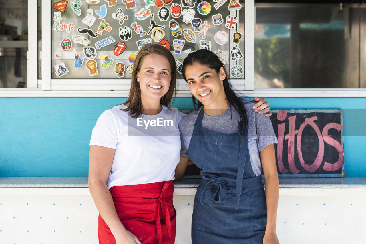 Portrait of confident young multi-ethnic female owners standing with arms around against food truck
