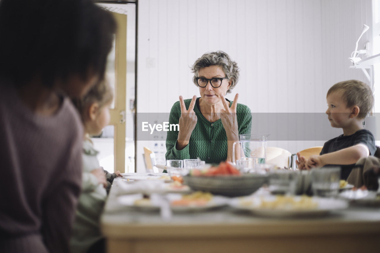 Senior female teacher communicating with students through hand signs while sitting at dining table during lunch break