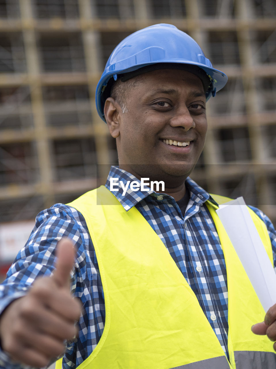 Portrait of an indian engineer holding rolled up plans, smiling at camera and showing a thumbs up