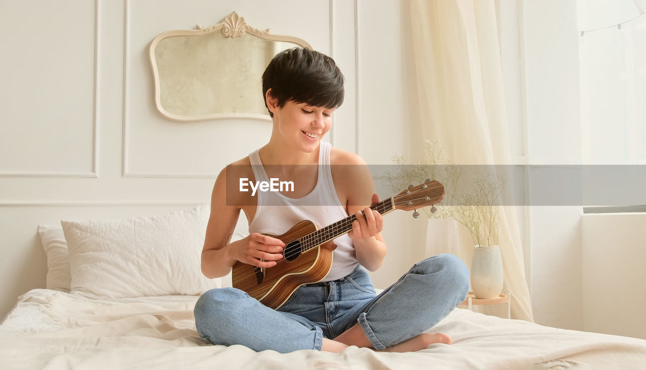 side view of young woman playing guitar while sitting on bed at home