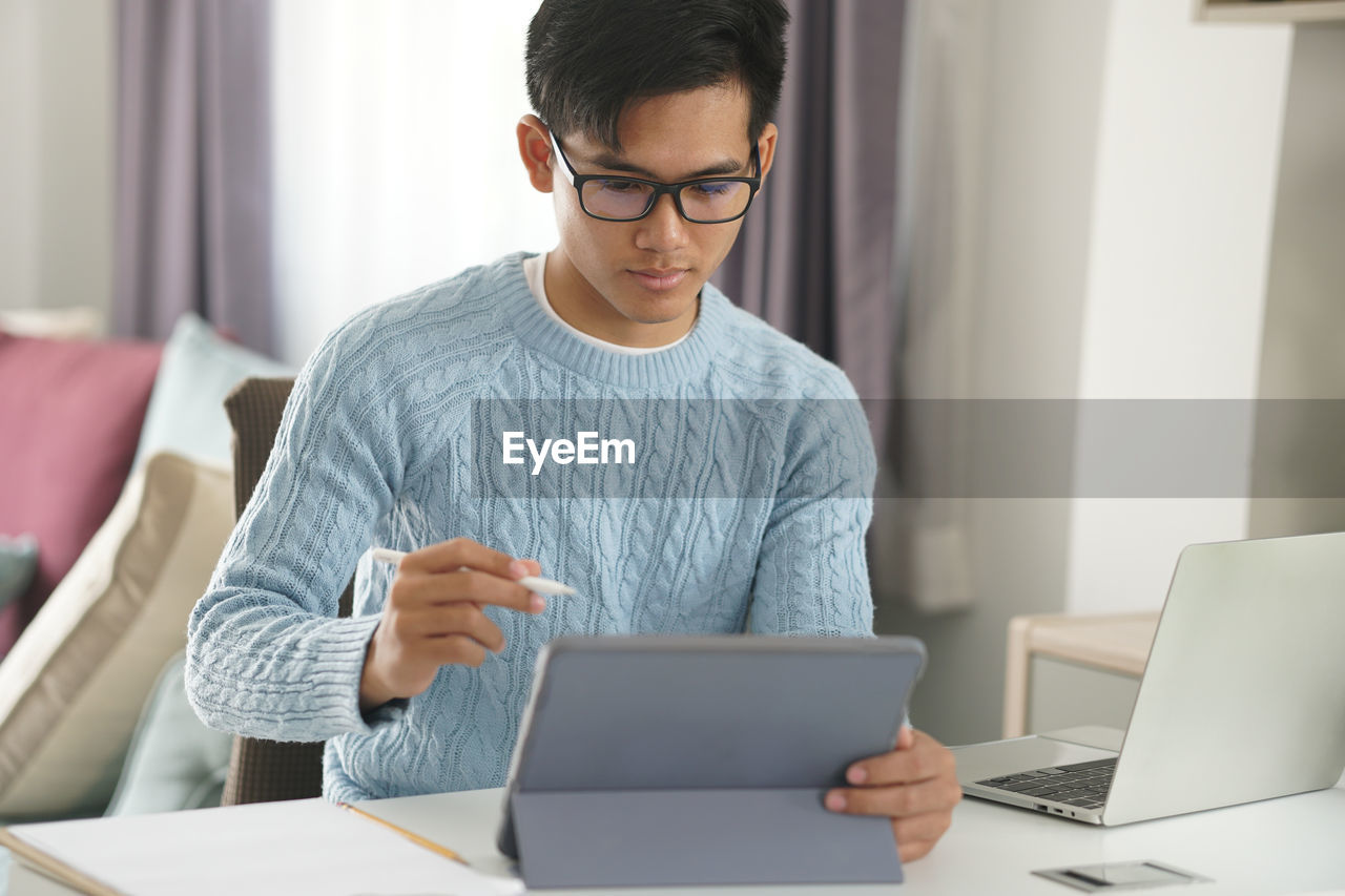 YOUNG MAN USING PHONE WHILE SITTING ON TABLE AT HOME