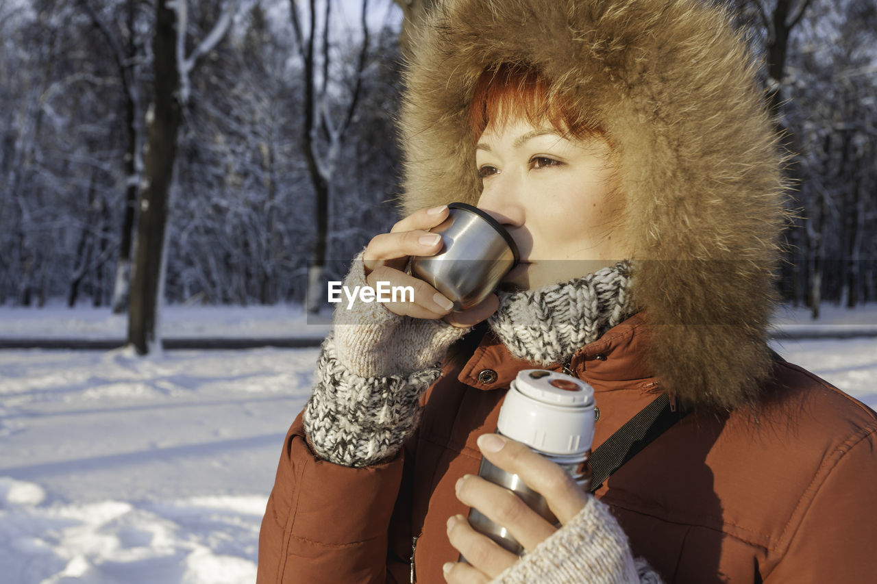 Woman in red coat drinks hot tea from metal thermos. leisure activity in winter forest at sunset.