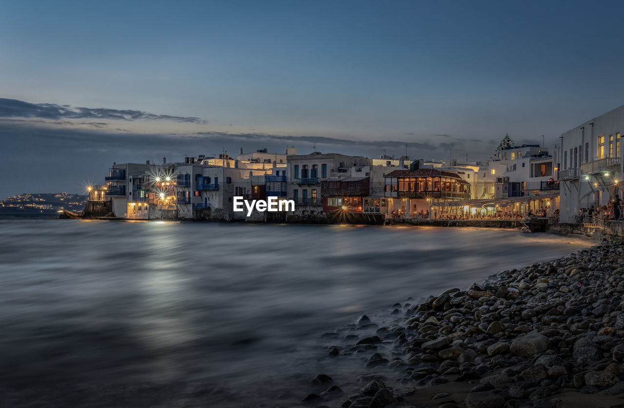 Illuminated buildings by sea against sky at dusk