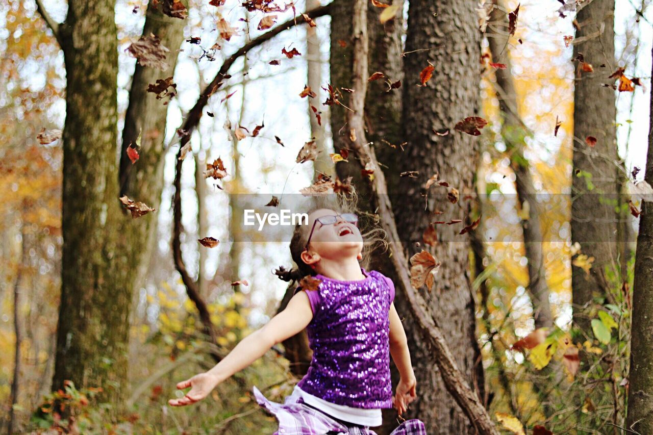 Girl throwing autumn leaves by bare trees in forest