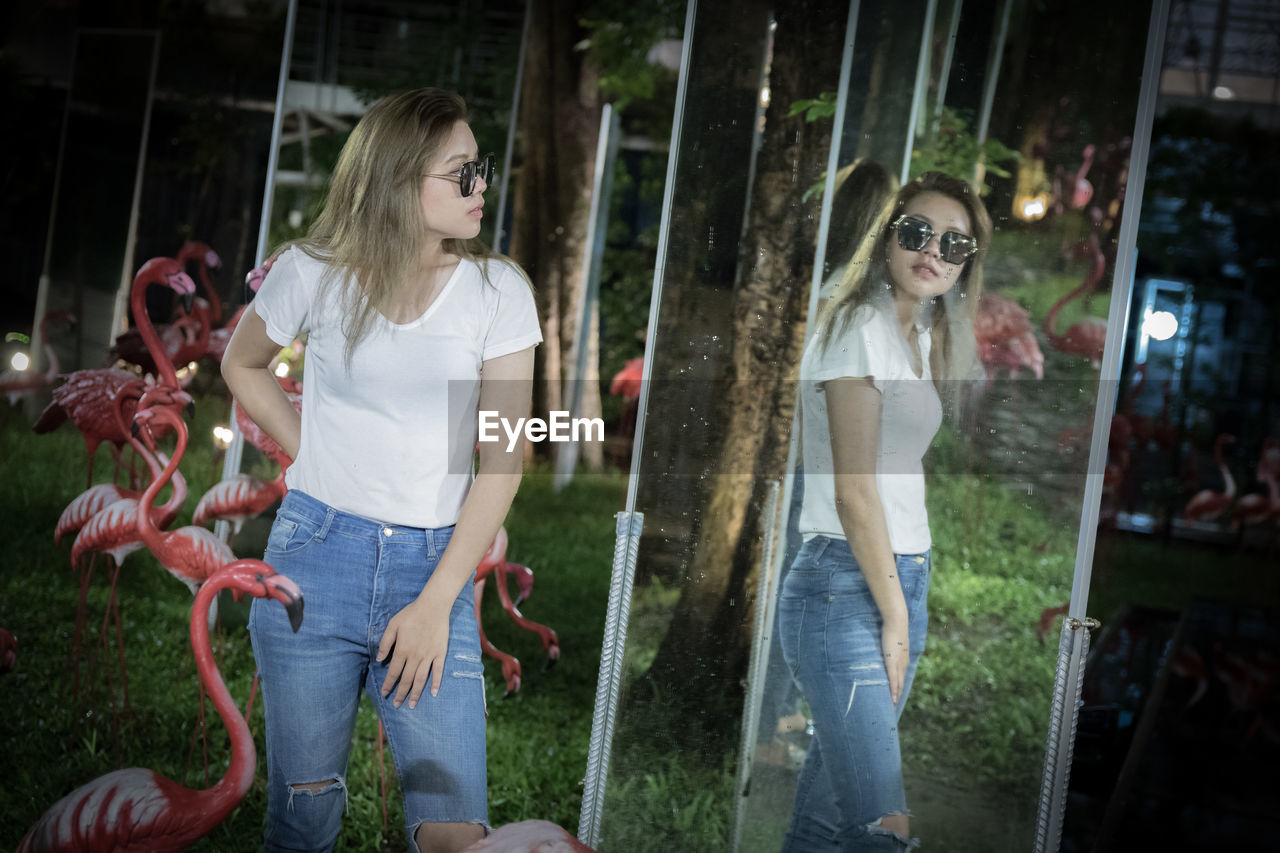 Young woman wearing sunglasses standing against mirror at night