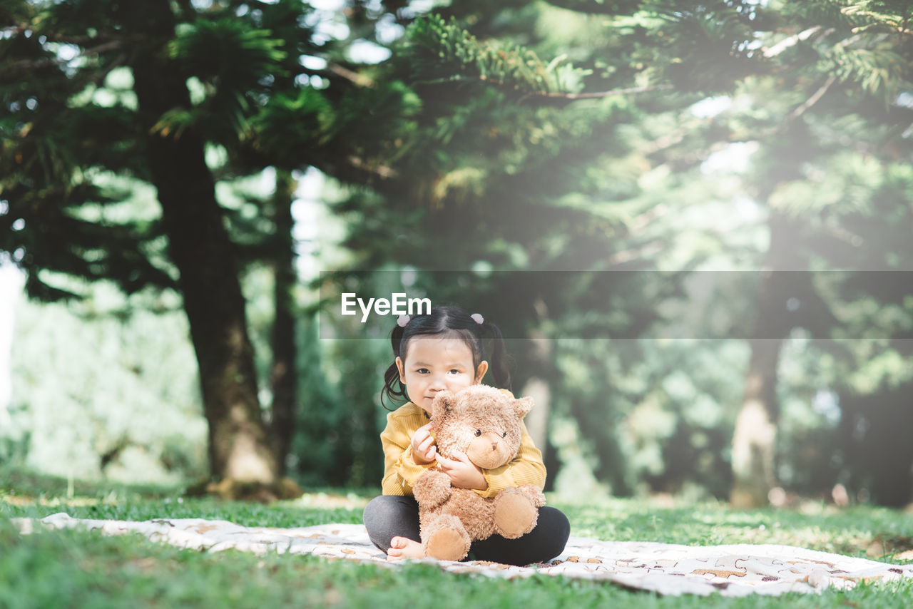 Portrait of cute girl sitting with toy on grass against trees