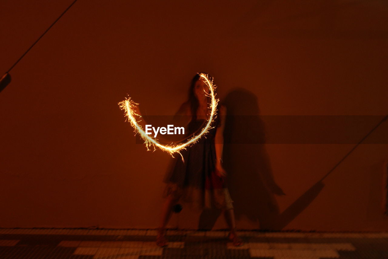 Blurred motion of woman holding illuminated sparkler against wall at night