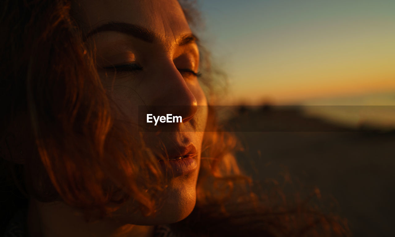Close-up of woman with closed eyes standing outdoors during sunset