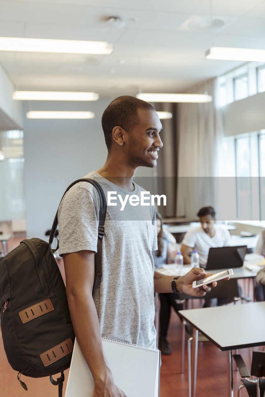 Smiling young student with backpack holding mobile phone while standing in classroom