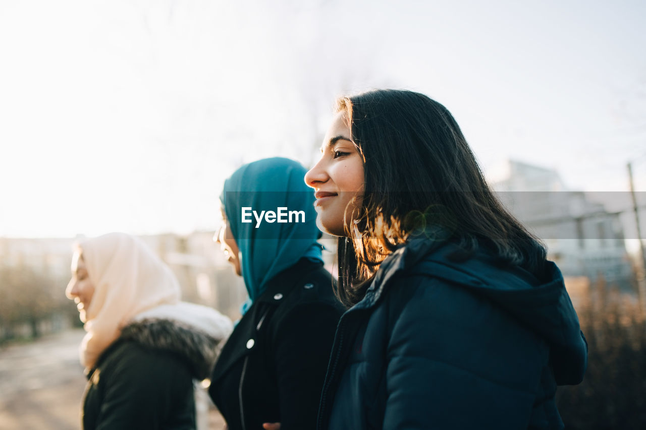 Side view of smiling teenage girl with young muslim friends in city against sky during sunset
