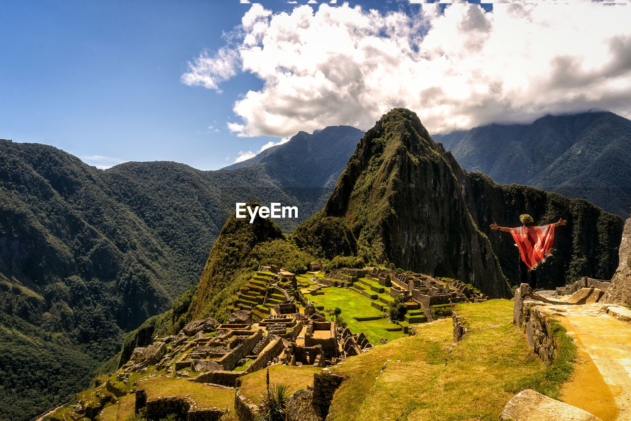 Back view of unrecognizable traveler in traditional poncho and hat admiring scenic mountainous valley while standing on viewpoint with outstretched arms during trip in machu picchu person