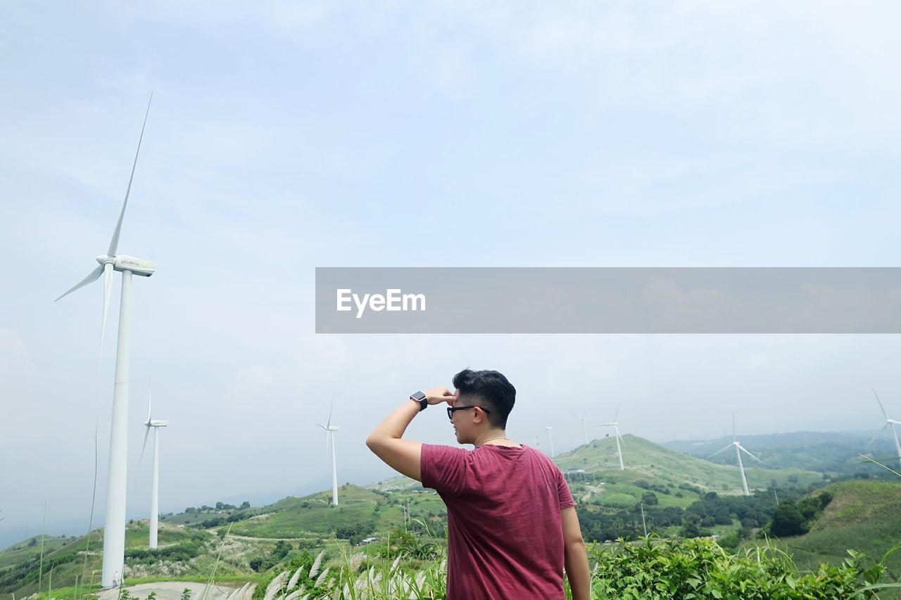 Man shielding eyes while looking away against windmills and sky on field