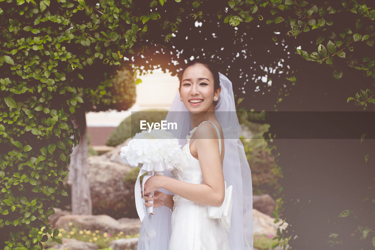 Portrait of smiling bride holding bouquet standing against trees in park