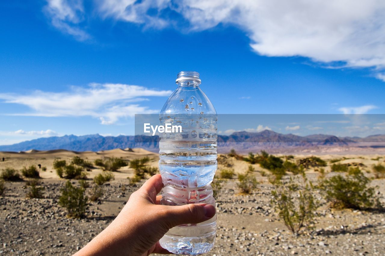 Cropped hand of person holding water bottle in desert against sky