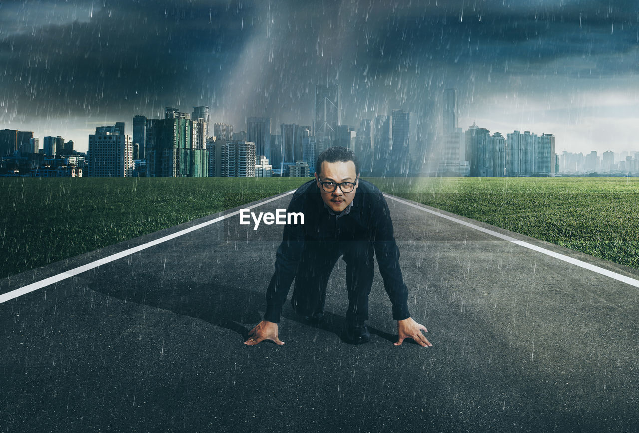 Portrait of businessman kneeling on road against cityscape during rainy season