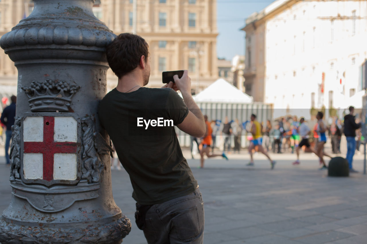 Rear view of man photographing athletes running during marathon in city