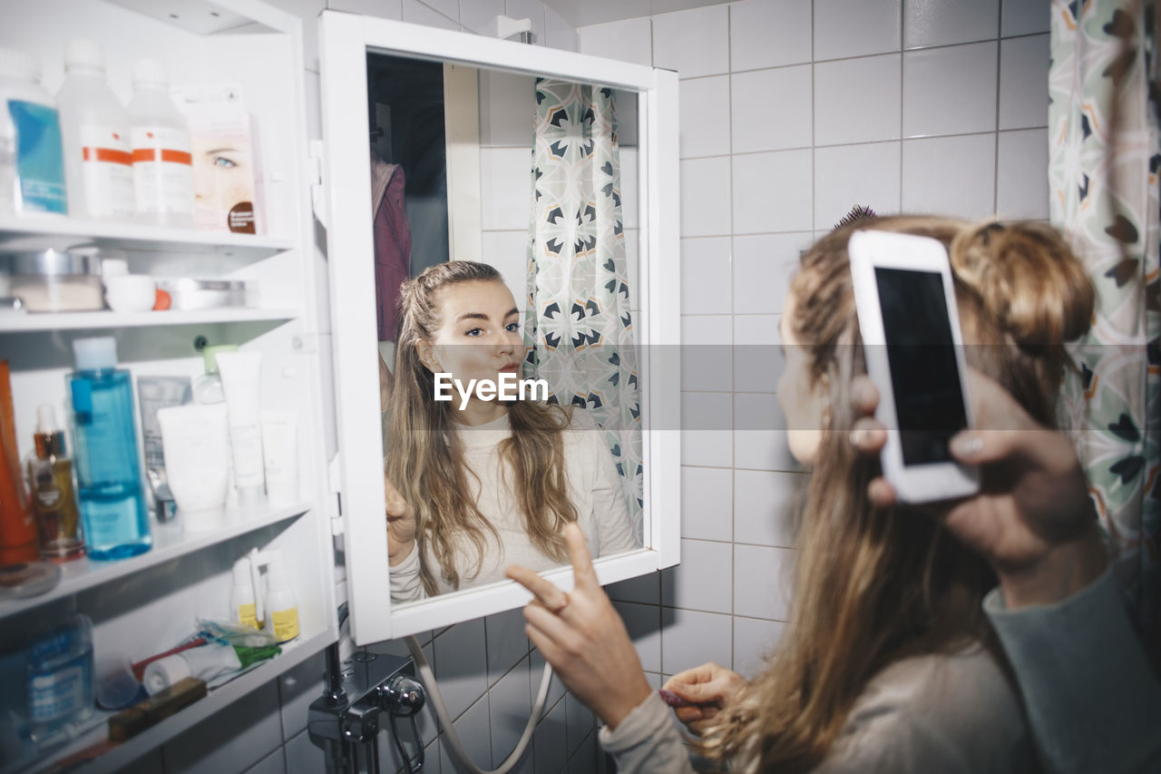 Cropped hand of woman photographing female friend in mirror reflection at dorm bathroom