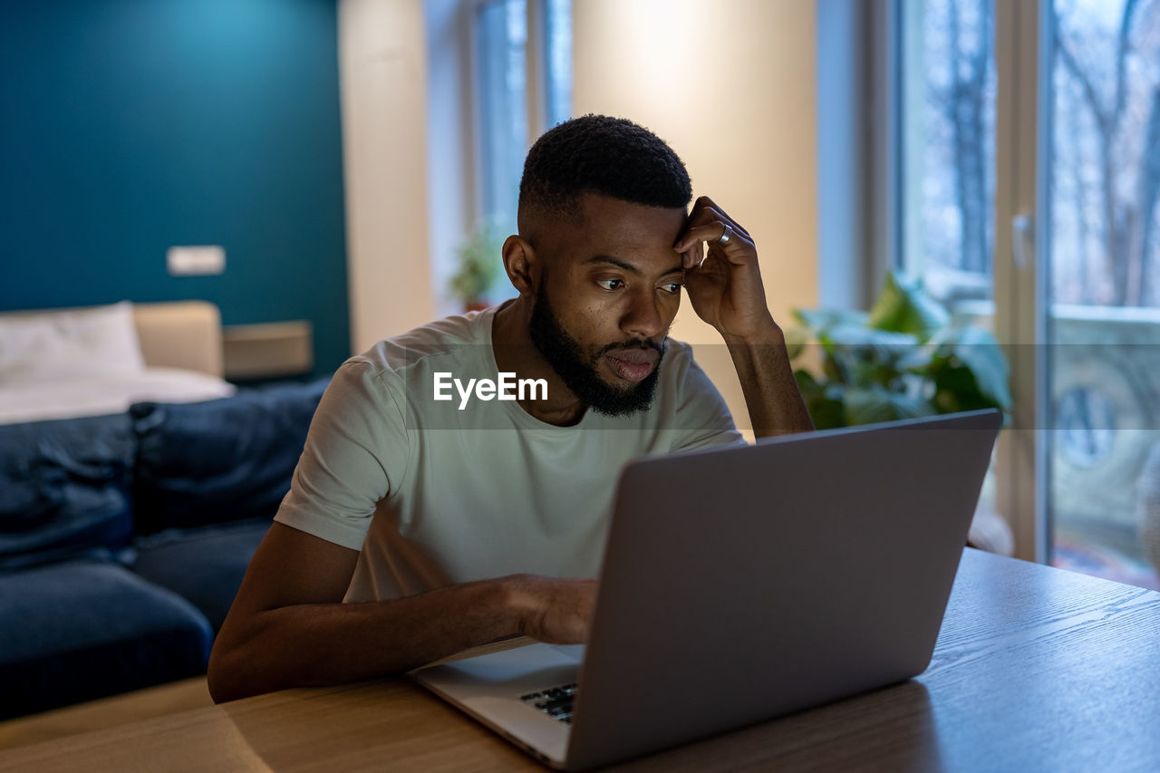 Focused african american guy freelancer working on laptop at home office, feeling overwhelmed